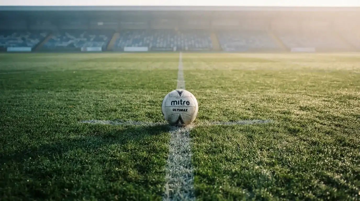 Ein einzelner Fussball liegt auf dem Anstosspunkt eines leeren Rasenplatzes am frühen Morgen, Tau glitzert auf dem Gras, die Tribüne liegt im Dunst.