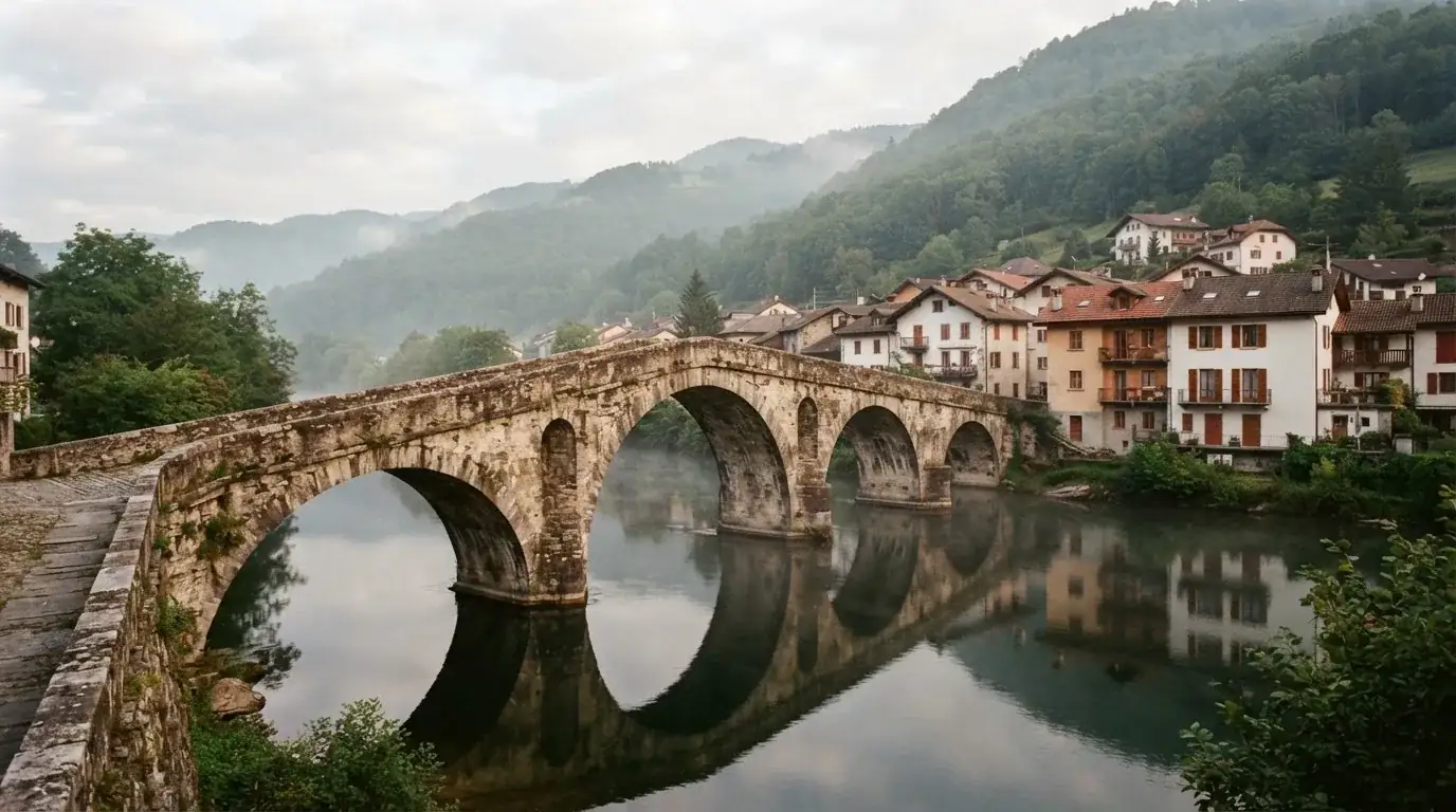 Eine alte Steinbrücke über einen ruhigen Fluss, dahinter Häuser mit Ziegeldächern und bewaldete Hügel.