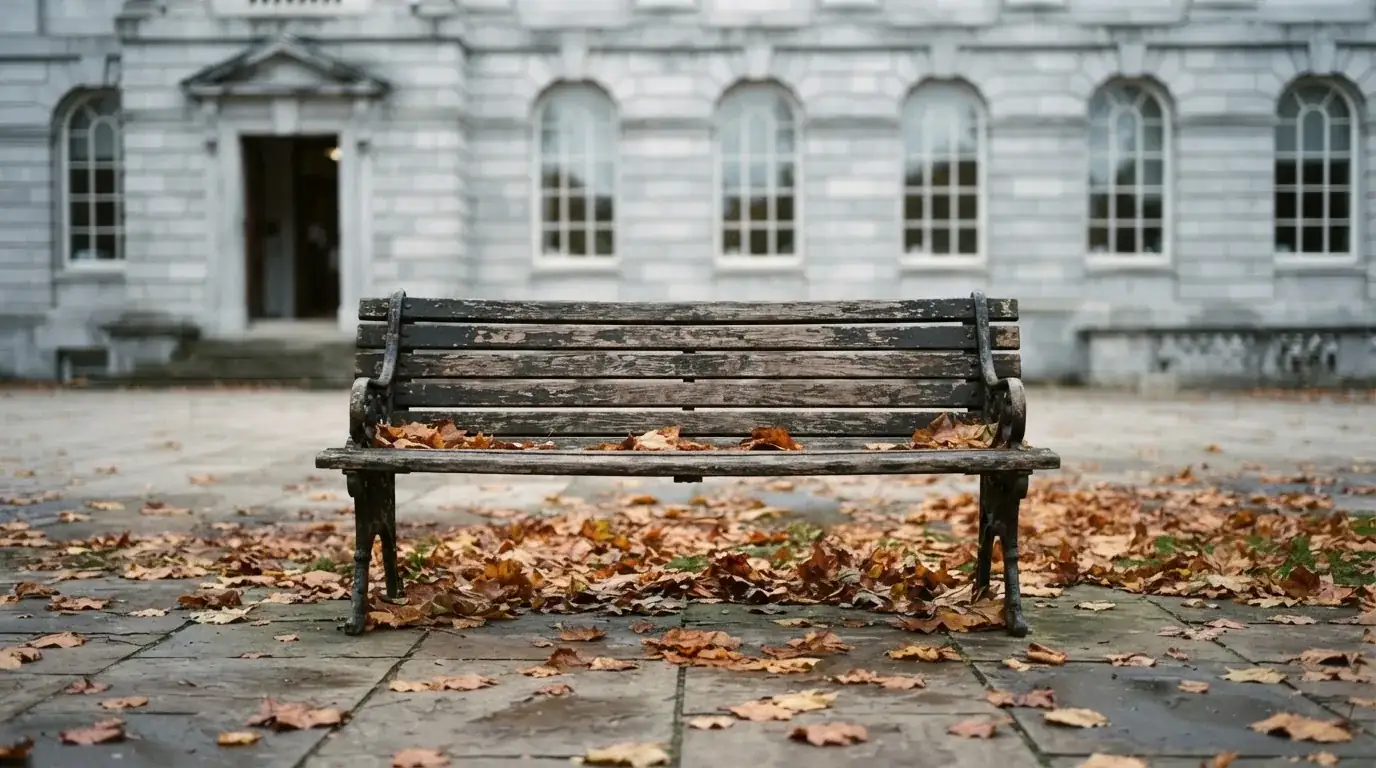 Eine Holzbank steht vor einem Verwaltungsgebäude aus Stein mit hohen Fenstern, Herbstlaub liegt auf dem Boden.