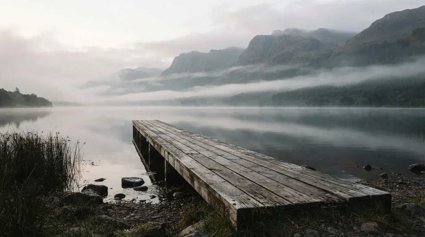 Ein Seeufer mit einem hölzernen Steg, Nebel liegt über dem Wasser, am anderen Ufer erheben sich Berge.