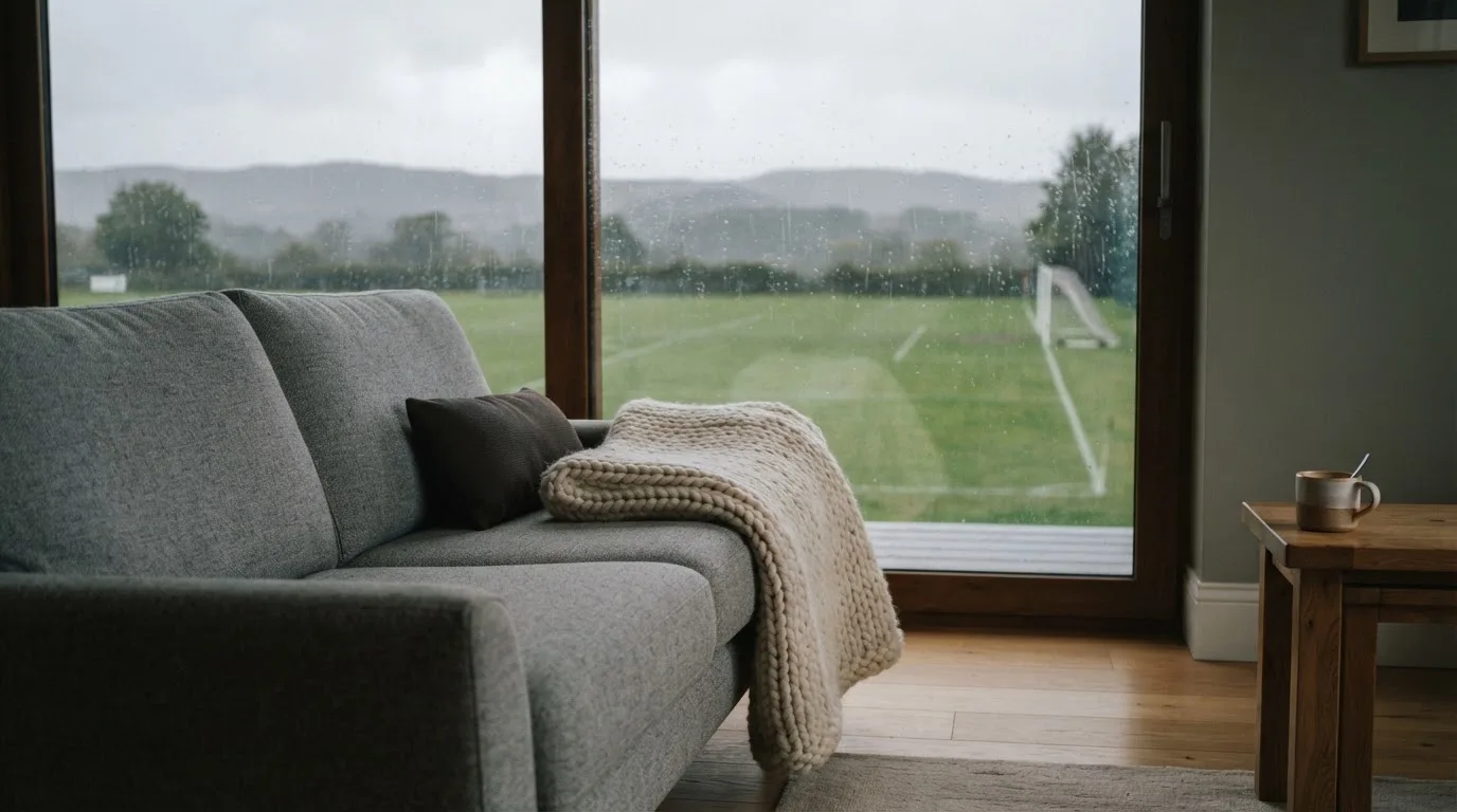 Ein Sofa mit Wolldecke steht vor einem grossen Fenster, draussen Regen und in der Ferne ein Fussballplatz.