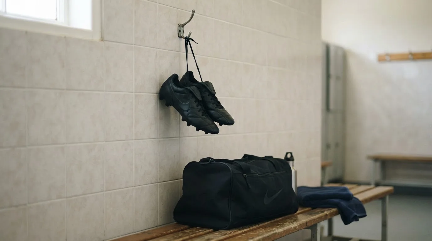Ein Paar Fussballschuhe hängt an einem Haken in einer ruhigen Umkleide, darunter steht eine Sporttasche auf einer Holzbank.