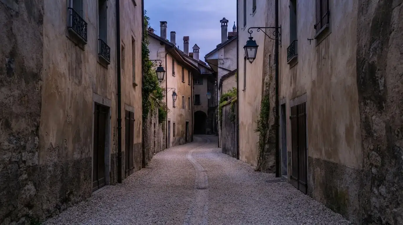 Ein schmaler Kiesweg führt zwischen zwei hellen Mauern durch eine Schweizer Altstadt, die Laternen sind noch nicht angezündet.