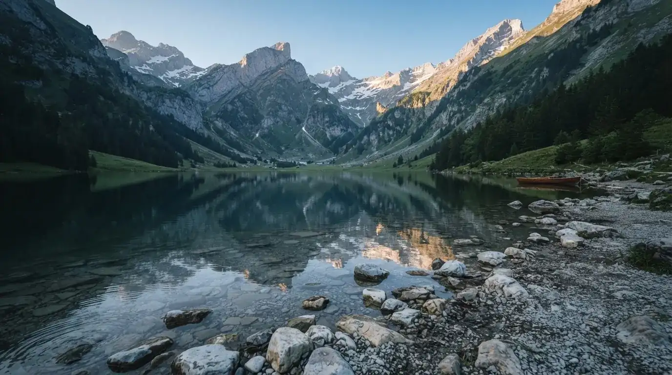 Ein Bergsee mit ruhigem Wasser und Gipfeln im Morgenlicht, am Ufer liegen glatte Steine.