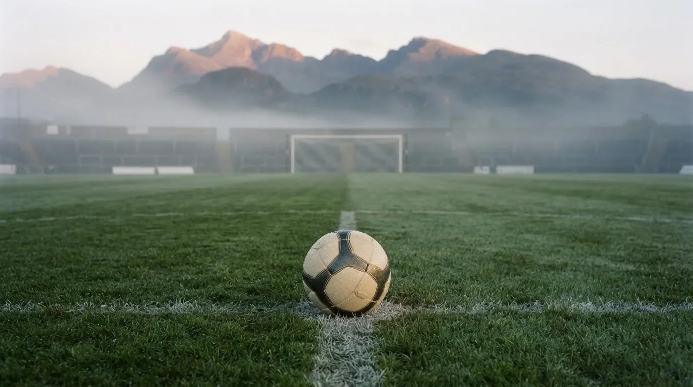 Ein einzelner Fussball auf dem Mittelpunkt eines leeren Rasens im Morgennebel, hinter dem Tor ragen Berge auf