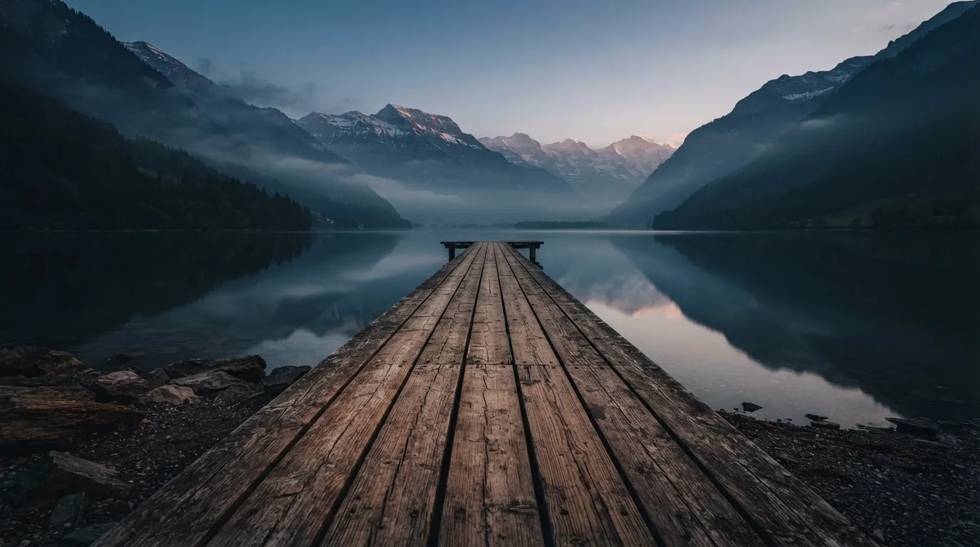 Ein Bergsee in der Dämmerung mit einem leeren Holzsteg, die Wasseroberfläche ist ruhig und Nebel zieht über die Hänge.