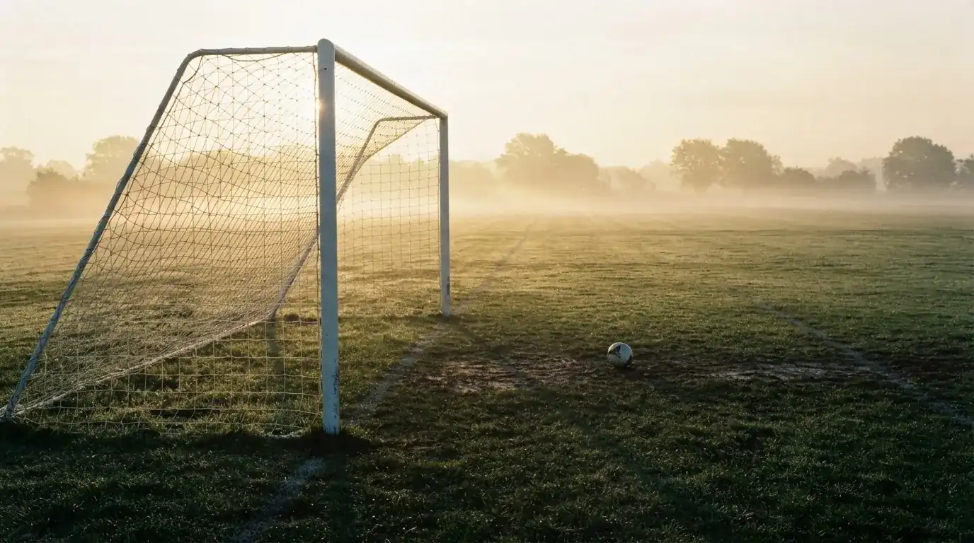 Ein Torpfosten mit Netz steht im Gegenlicht auf einem leeren Fussballplatz, über dem Rasen liegt Nebel.
